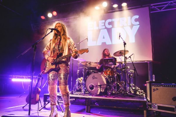 Holly Rankin performing with Jack River at Electric Lady, wearing a glittering suit and playing guitar in front of the drum riser and a projection of the event's title.